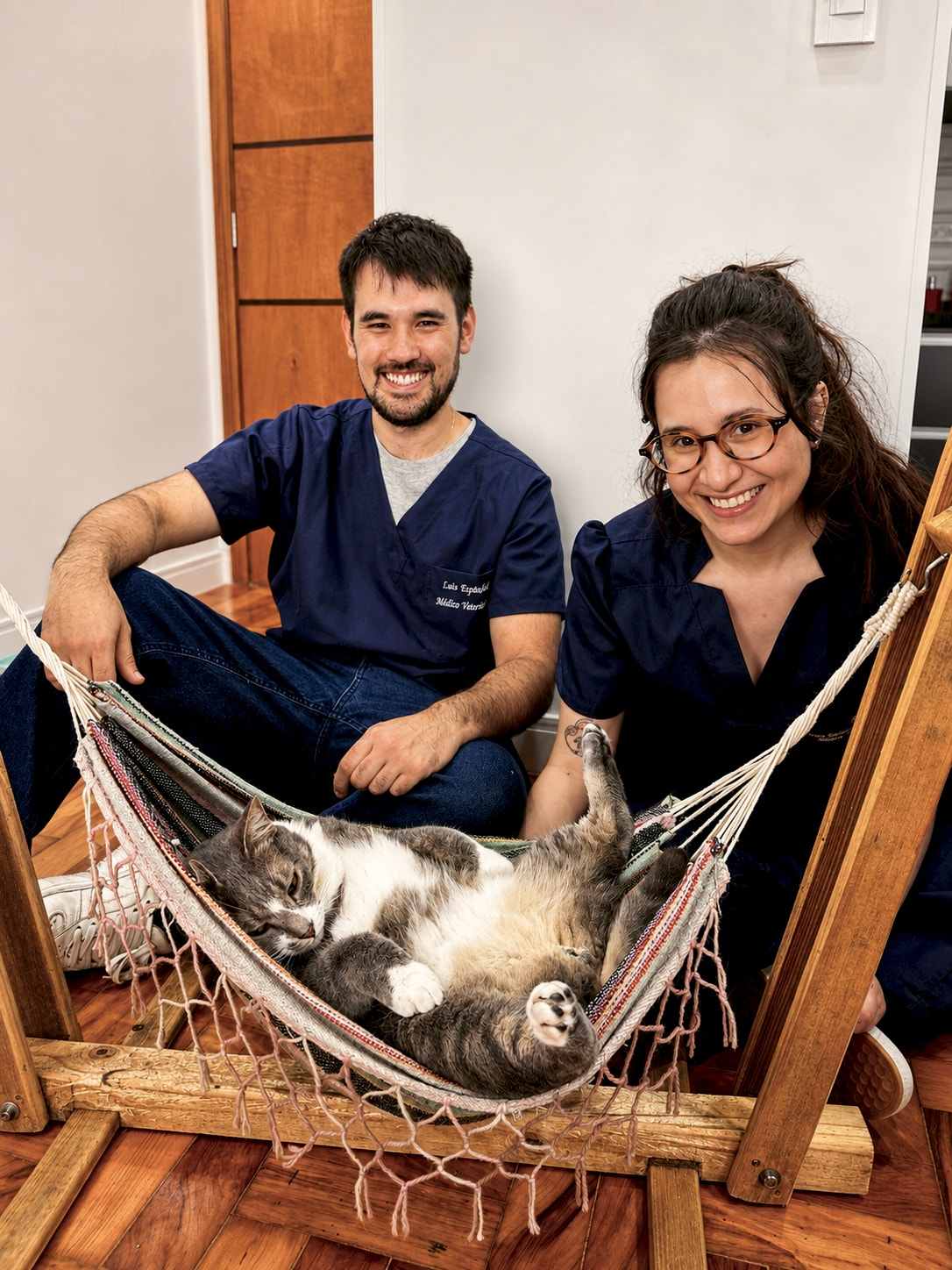 Barbara e Luiz sentados no chão, sorrindo, ao lado de um gato cinza que dorme de barriga para cima na rede